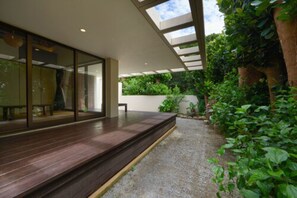 A modern outdoor patio with a wooden deck, glass walls, and a view of lush greenery. - Private Villas Ryuz BISE (Motobu)