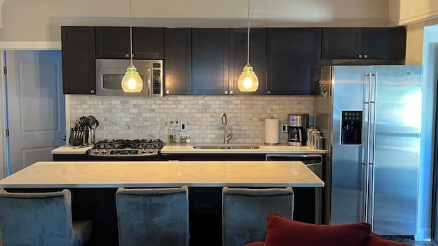 Une cuisine moderne avec des meubles de couleur sombre, un réfrigérateur en acier inoxydable, un four encastré, une machine à café et des luminaires suspendus.