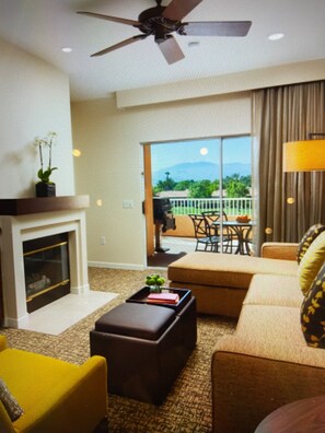 Cozy living room with a fireplace, ceiling fan, and large window offering a view of the outdoors.
