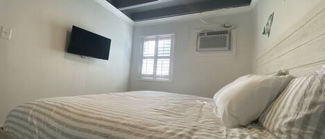 A bedroom with a bed, a mounted TV, a window with blinds, and an air conditioning unit.