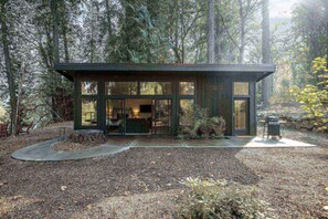 A modern cabin with large windows, a visible kitchen area, and a small outdoor seating area.
