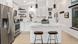 A modern kitchen with white cabinetry, stainless steel appliances, a marble island with stools, pendant lighting, and wall art. - Panoramic Riverside and Lush Forests Views, Pet Friendly, New Décor, Steps Away From Dockside (West Linn)