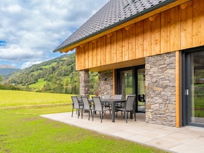 Een modern gebouw met een gevel van natuursteen en een houten gevel, met een terras, zitplaatsen buiten en uitzicht op een bergachtig landschap.