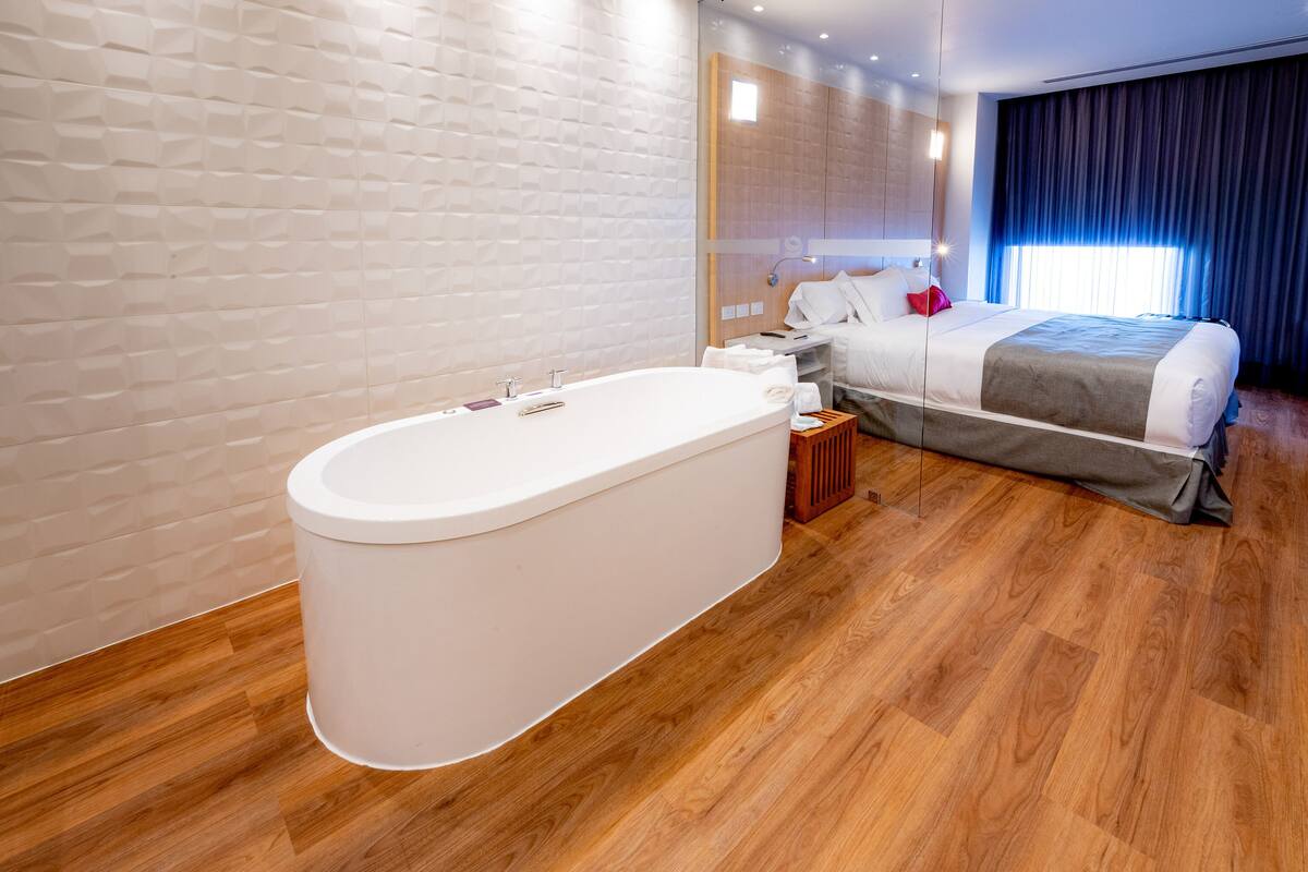 Une chambre d’hôtel moderne dotée d’une grande baignoire, d’un plancher en bois, d’un grand lit avec une couvre-lit grise et blanche, et d’une grande fenêtre avec des rideaux.