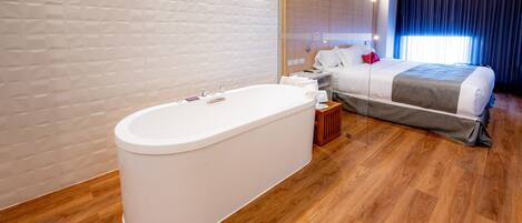 Una habitación de hotel moderna con una bañera grande, suelo de madera, una cama grande con colcha gris y blanca, y un ventanal con cortinas.