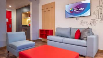 A modern hotel lobby with a grey sofa, a red ottoman, and a television displaying 'Ramada Encore by Wyndham'.