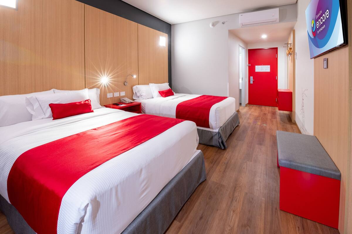 Une chambre d’hôtel avec deux lits, une décoration aux couleurs rouge et blanche, des panneaux de bois et un téléviseur fixé au mur.