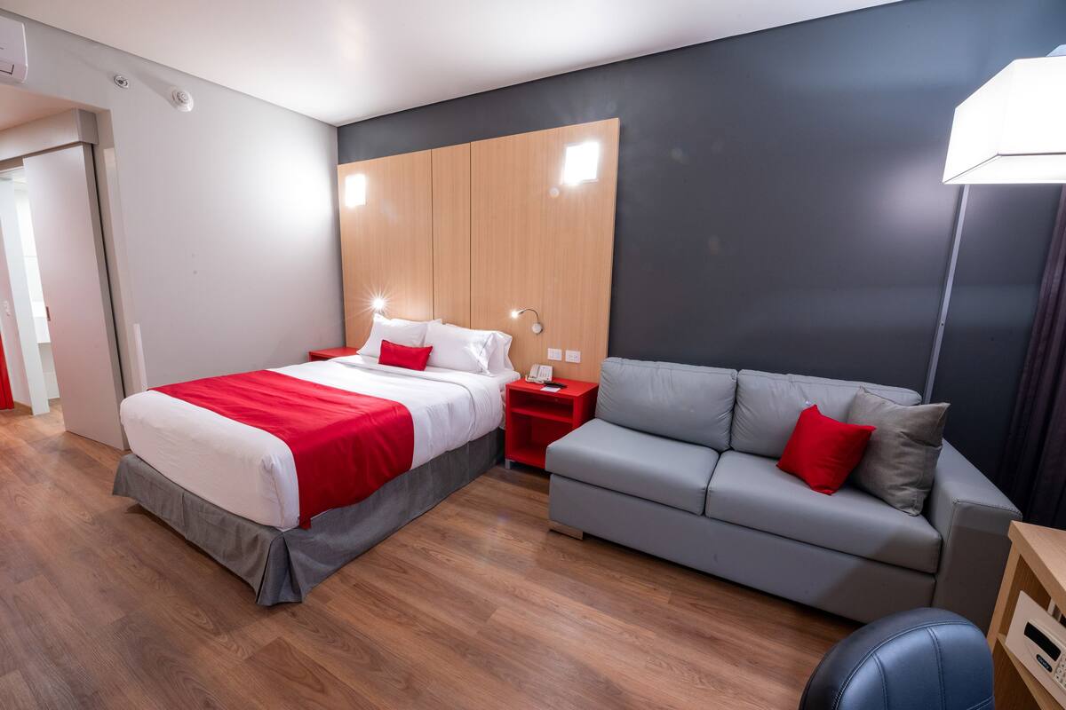 Une chambre d’hôtel moderne avec un lit, un canapé gris, une tête de lit en bois et une table de chevet en bois.