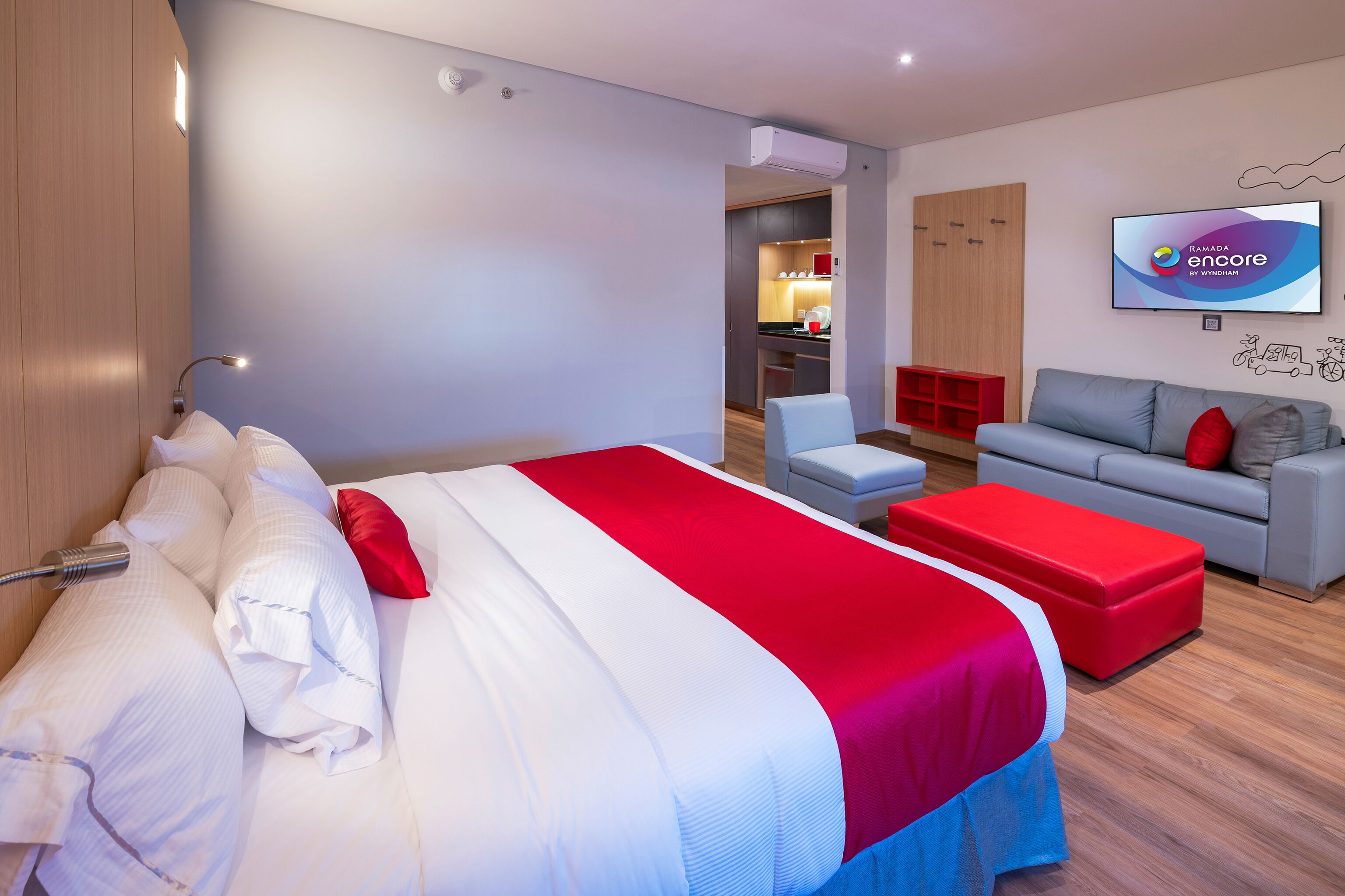 A modern hotel room with a large bed, a grey sofa, a red ottoman, and a TV displaying 'Ramada encore by Wyndham'.