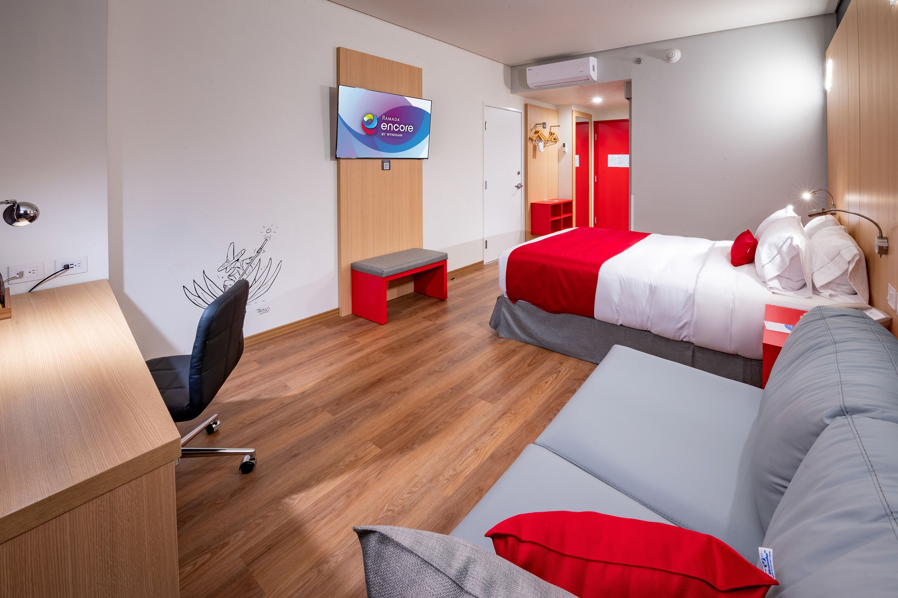 A modern hotel room with a wooden desk, a TV, a bed with red bedding, a grey sofa, and a red bench.