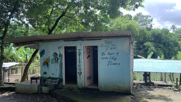 Un petit bâtiment rustique, doté d’un toit en tôle ondulée, d’une porte portant l’inscription « Q » et de graffitis sur les murs.