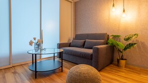 A modern living room with a grey sofa, a round coffee table, a woven pouf, and a potted plant. - Exia Daikoku AFP Apartment (Osaka)