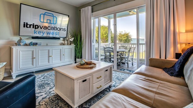Ein Wohnzimmer mit einem Fernseher, der 'itripvacations Key West' zeigt, einem weißen Schrank, einem Couchtisch, einer Couch und Blick ins Freie durch große Schiebetüren.