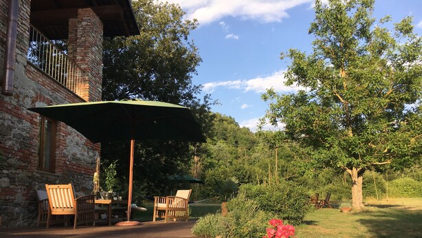 A rustic outdoor seating area with a brick building, wooden chairs, a table, and a green umbrella. - Casa al Tanaro | Spacious country house with pool in southern Piedmont (Castellino Tanaro)