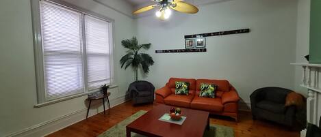 A living room with a wooden floor, a ceiling fan, a sofa with tropical cushions, a small table with a fruit bowl, and a potted plant.