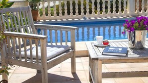 Un espace aménagé au bord de la piscine, avec une chaise en bois, une table sur laquelle se trouve un livre et un vase de fleurs, et une vue sur la piscine et les bâtiments en arrière-plan.