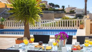 Un espace repas aménagé au bord de la piscine, avec des tables préparées pour le petit-déjeuner, comprenant des croissants et du jus d’orange.