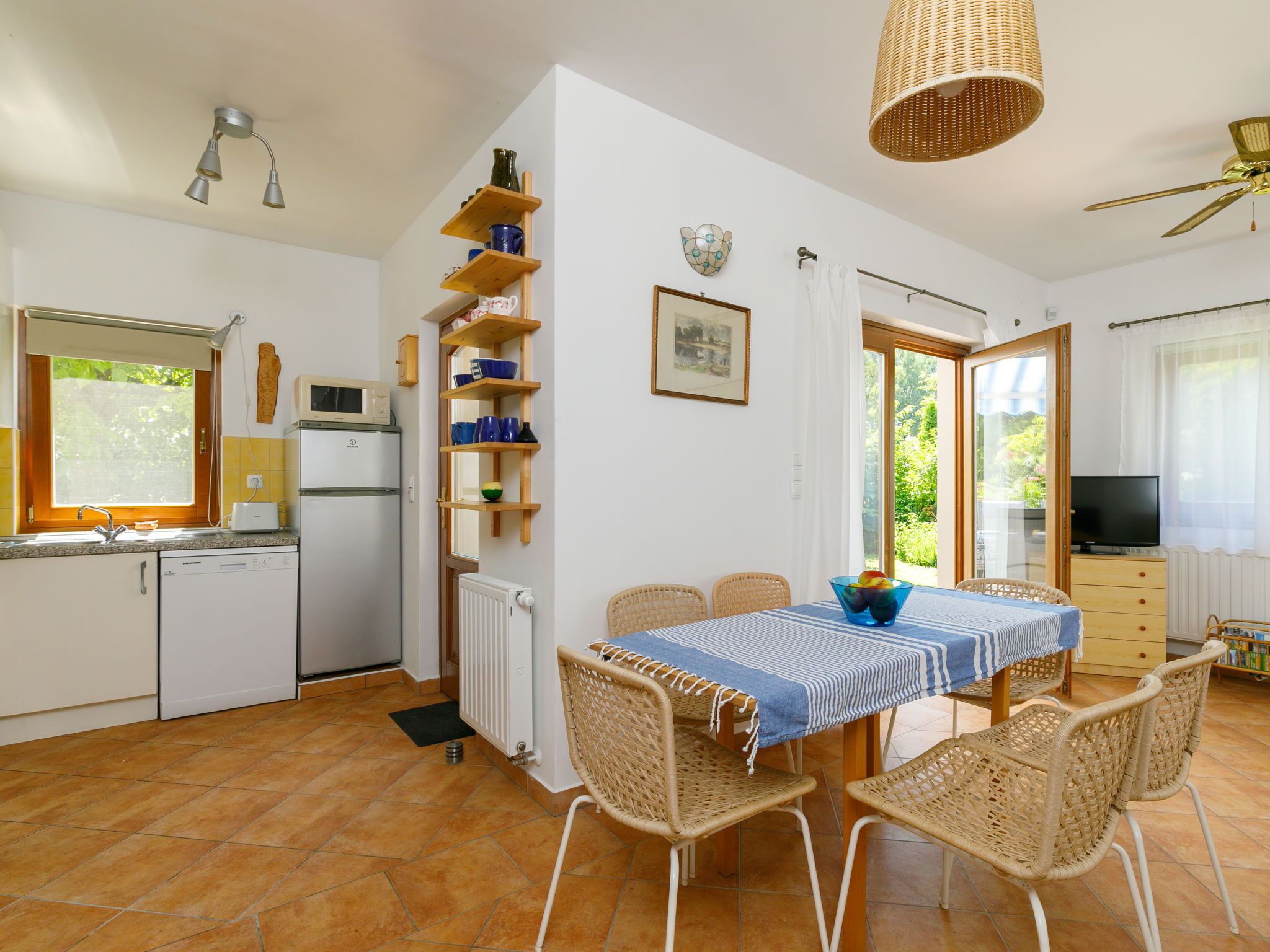 Cocina compacta con armarios blancos, microondas integrado y refrigerador pequeño. Cuenta con una zona de comedor con mesa y sillas de mimbre, además de un ventanal que proporciona luz natural.