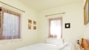 Une chambre à coucher avec un lit, deux fenêtres avec des rideaux, un radiateur et une table de chevet avec une lampe.
