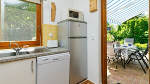 Une cuisine compacte équipée d’un lave-vaisselle, d’un four à micro-ondes et d’un réfrigérateur. Une fenêtre laisse entrer la lumière naturelle et offre une vue sur le jardin.