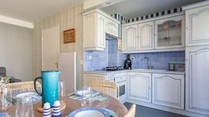 A dining area with a round table set for four, surrounded by wicker chairs. A kitchen with white cabinets and blue tiles is visible in the background. - Montaigu by Interhome (Cabourg)