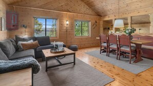 Een knus interieur van een chalet met houten plafond, een bank, een eettafel met stoelen en grote ramen met uitzicht op de omgeving.