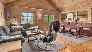 Een gezellig interieur van een blokhut met een houten plafond, een bank, een schommelstoel, een eettafel met stoelen en uitzicht op de natuur door grote ramen.