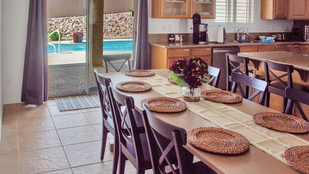 Comedor con mesa lista para cuatro, cocina al fondo y vista a una piscina a través de un ventanal.