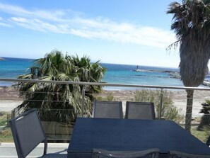 En balkon med et bord og stole, udsigt over en strand med palmer og et fyrtårn.