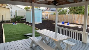 A covered outdoor patio with a picnic table and benches, surrounded by a wooden deck and a fenced backyard with a lawn and a shed.