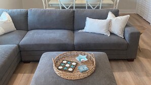 A living room with a grey sectional sofa, a matching ottoman, and a wicker tray with decorative items.