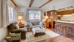 A cozy living room with a plaid sofa, a coffee table, and a bookshelf. A kitchen area with wooden cabinets and a countertop is visible. - Rocky Mountain Riverside Escape; 1 Mile to Park (Estes Park)