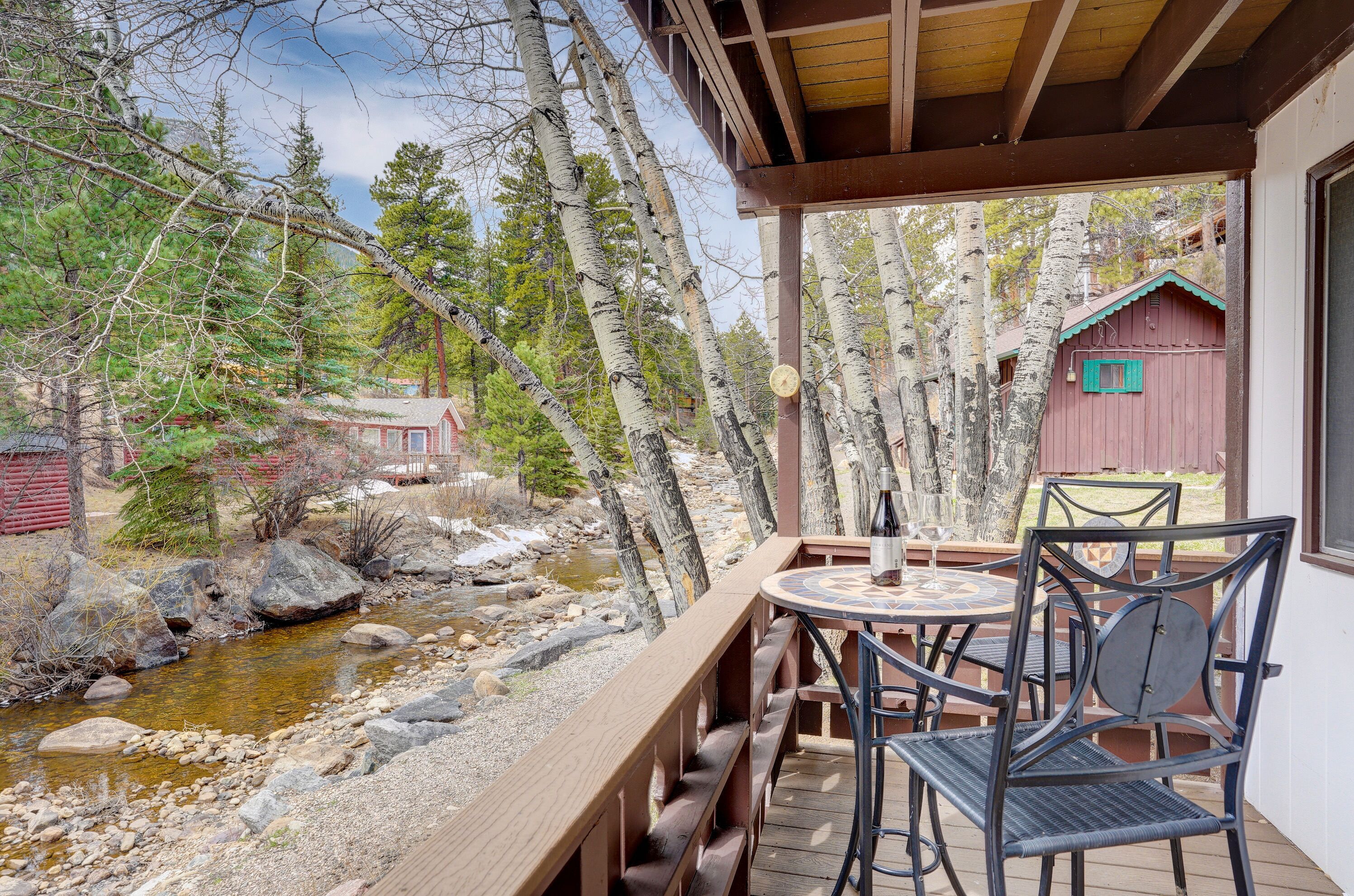 Eine Holzterrasse mit Tisch und Stühlen mit Blick auf einen Bach und ein rotes Gebäude.