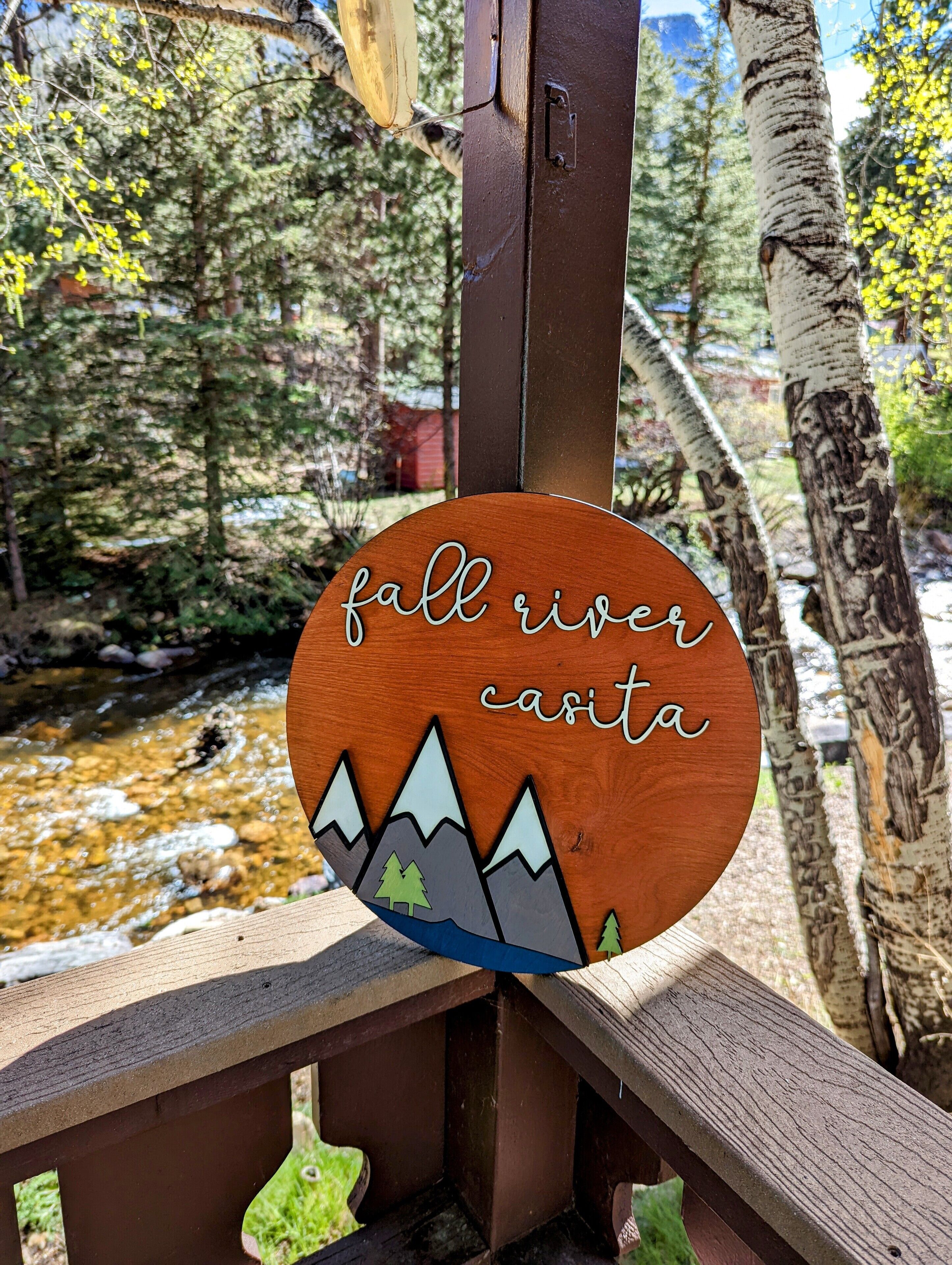 A wooden sign with "fall river casita" text and mountain graphics, set against a backdrop of trees and a stream.