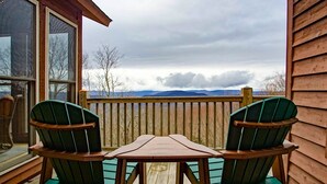 Una terraza de madera con dos sillas verdes y una mesita, con vistas a un paisaje montañoso.