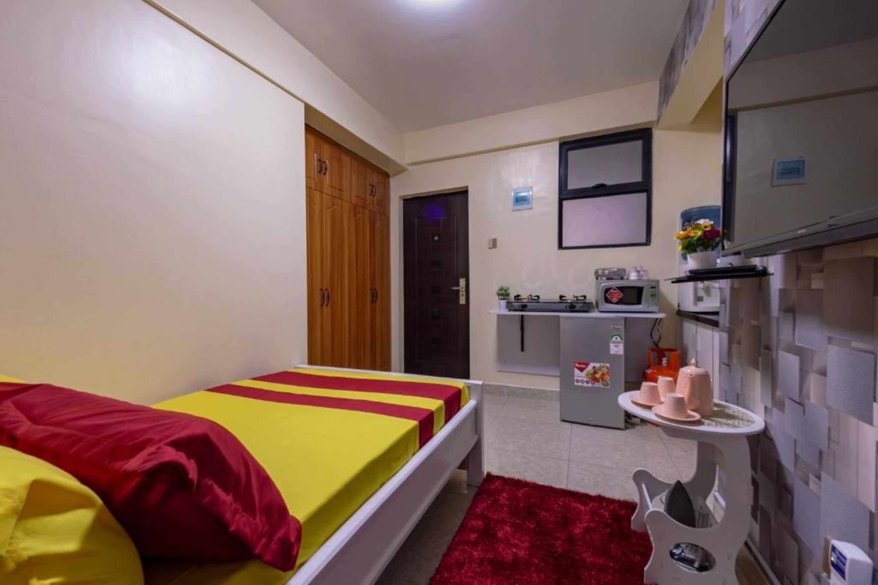 A compact room with a bed, a kitchenette, and a small table with items.