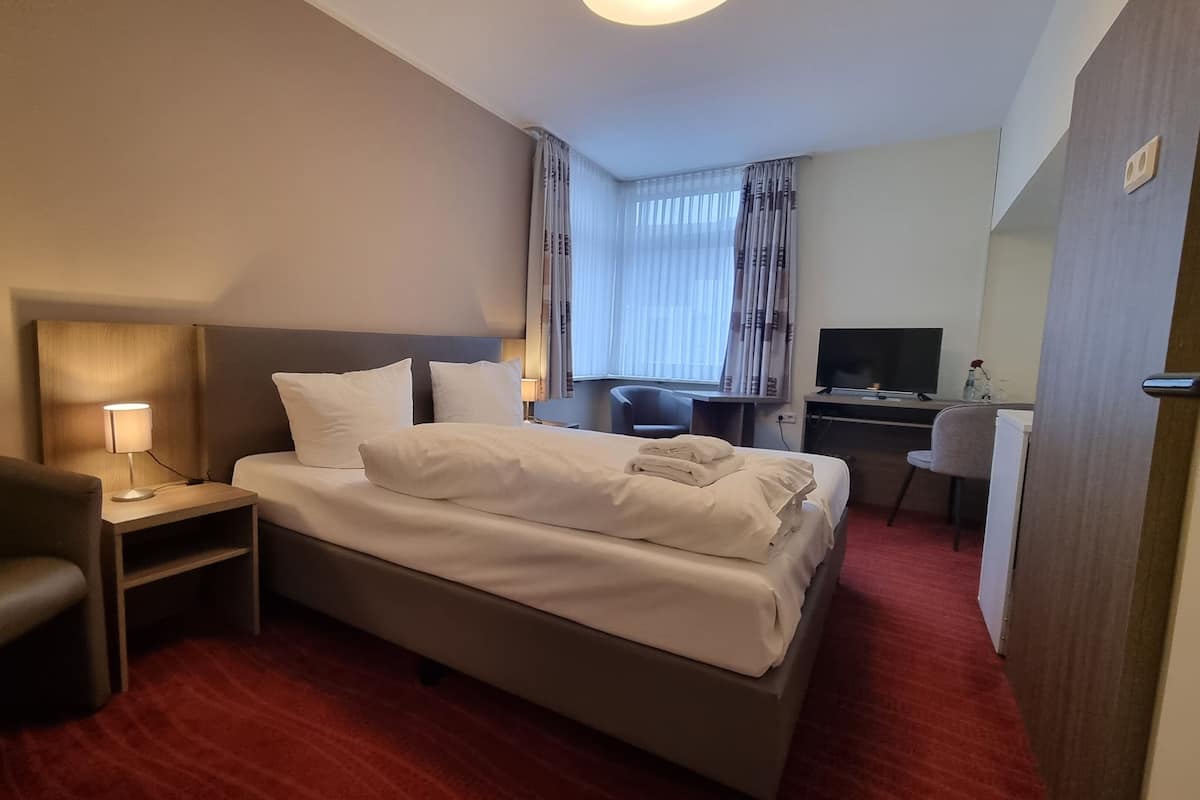 A hotel room with a bed, a desk with a computer, a chair, and a lamp.