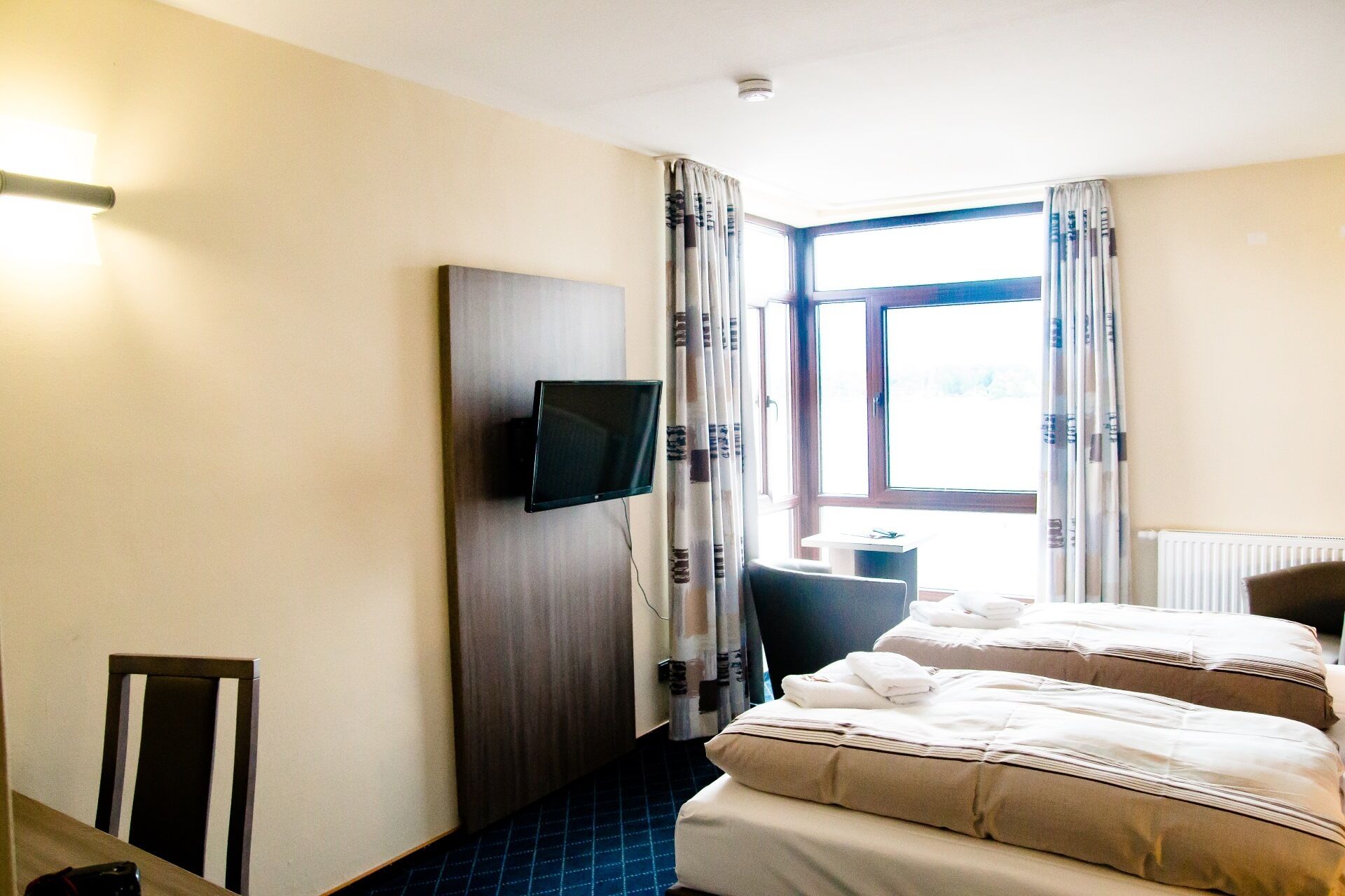 Ein Hotelzimmer mit einem Bett, einem an der Wand befestigten Fernseher, einem Sessel und einem Fenster mit Vorhängen.