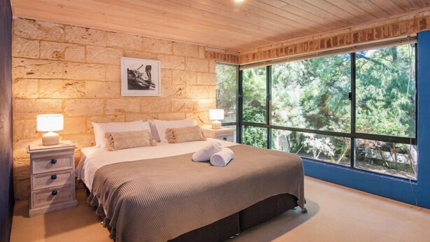 Un dormitorio con pared de piedra, un ventanal con vistas a árboles, una cama con manta marrón y dos lámparas de noche.