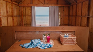 Une cabane en bois avec vue sur la plage. Elle comprend un banc, une table avec un panier et une bouteille, ainsi qu’une fenêtre avec des rideaux.