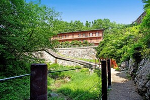 Un edificio de varios pisos con balcones, rodeado de abundante vegetación y un puente de piedra.