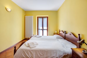 A hotel room with two beds, a radiator, a window with curtains, a bedside table with a lamp, and a wall-mounted light fixture.