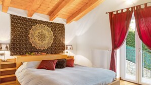 Un dormitori amb un llit, capçalera de fusta, un teixit mural i vista a través d'una finestra amb cortines.