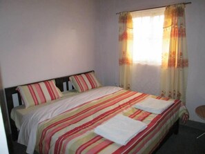 Une chambre avec un lit simple, une couvre-lit à rayures, une tête de lit sobre et une fenêtre ornée de rideaux à motifs floraux.