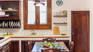 A kitchen with a wooden cabinet, a window with lace curtains, a tiled backsplash, and a dining table with chairs. - Villa 'Brezza di Limoni' close to the Sea with Terraces & A/C (San Vito lo capo)