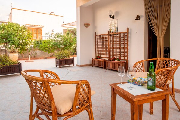 A patio with wicker furniture, a table with a bottle and glasses, and a view of a building with a tiled roof.