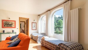 A room with a sofa, two beds with plaid bedding, a radiator, and a window with curtains.