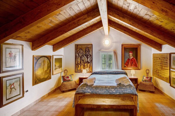 A bedroom with a wooden ceiling, a bed, two nightstands, a window, and various framed artworks on the walls.