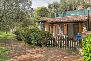Une maison de campagne rustique, avec un chemin de briques menant à un portail en bois, entourée d’une végétation luxuriante et d’un mur de pierre.