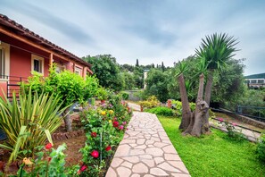 A stone pathway leading to a building with a red facade, surrounded by lush greenery and flowering plants.